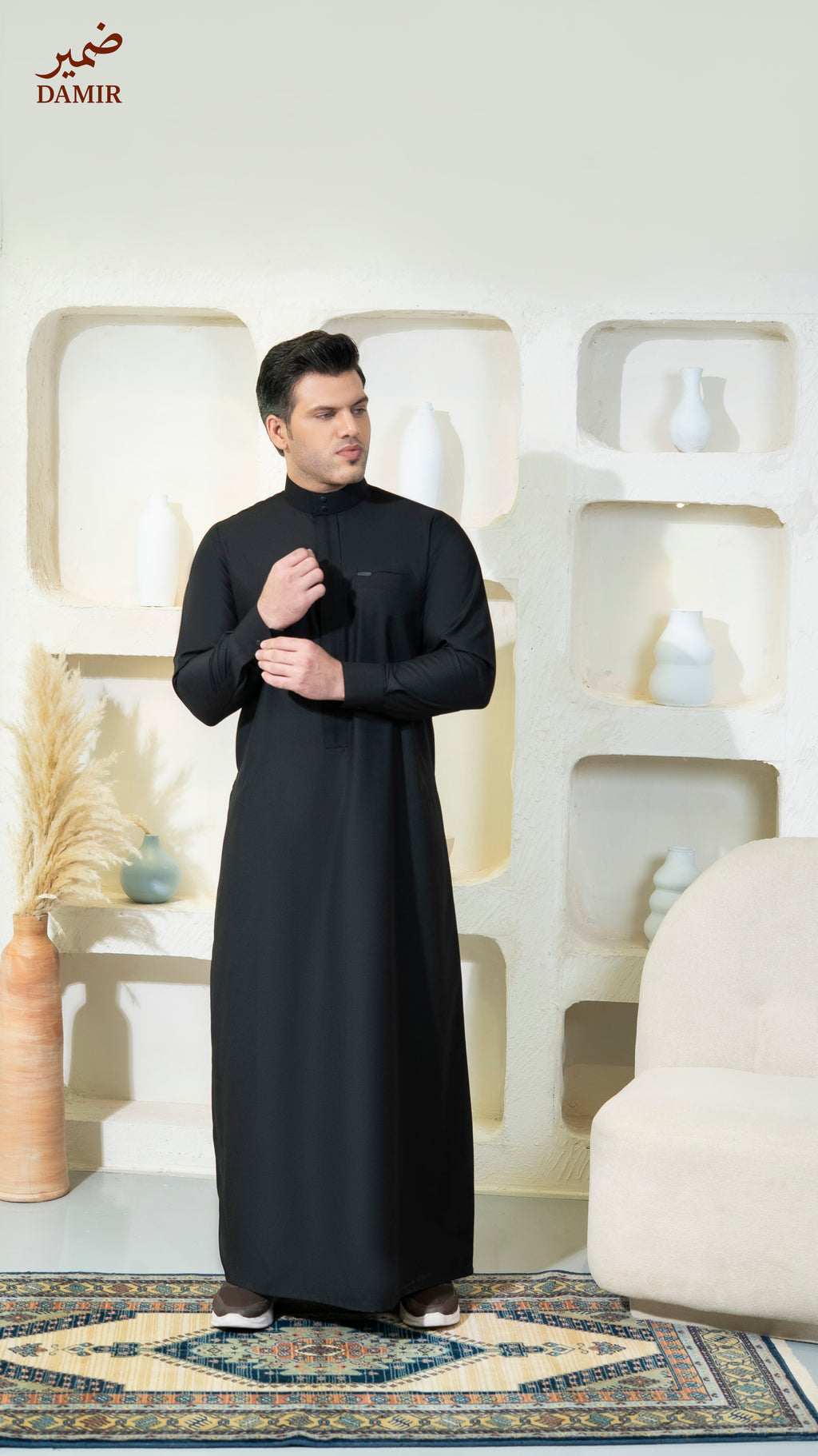 Man wearing a black traditional outfit in a decorative room with 'DAMIR' branding.