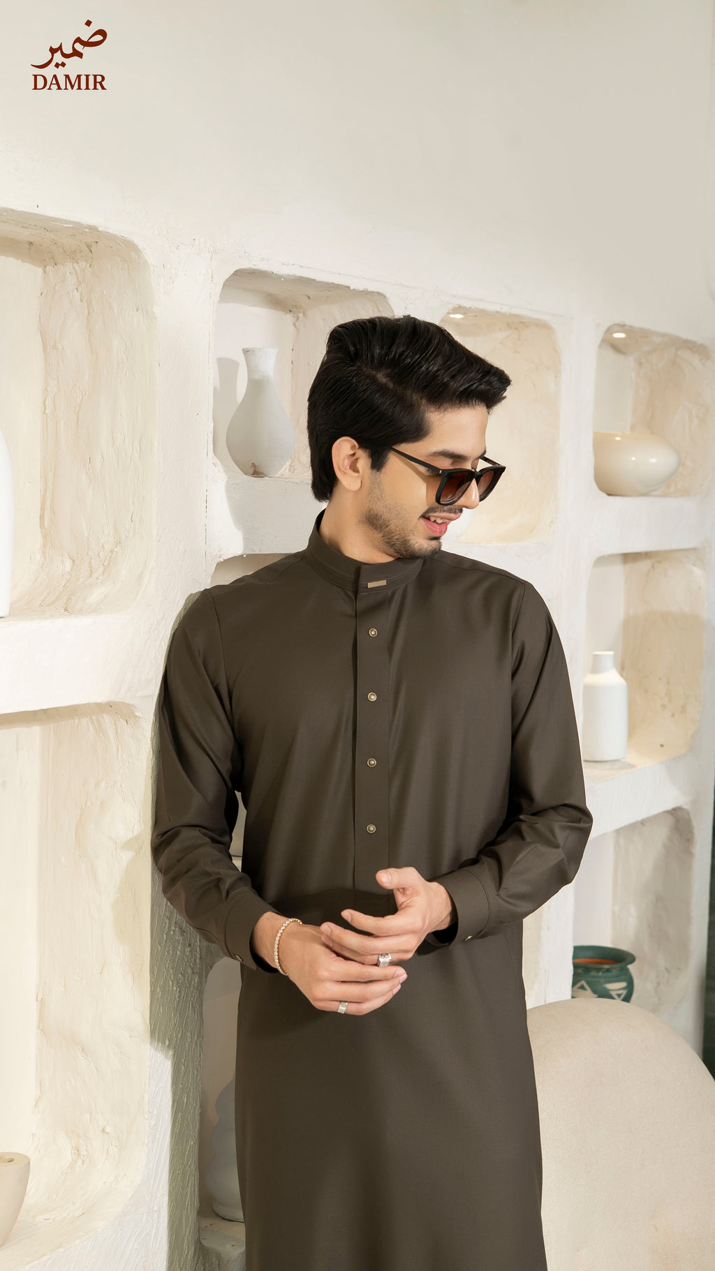 Man wearing a dark brown traditional outfit standing against a white decorative wall.