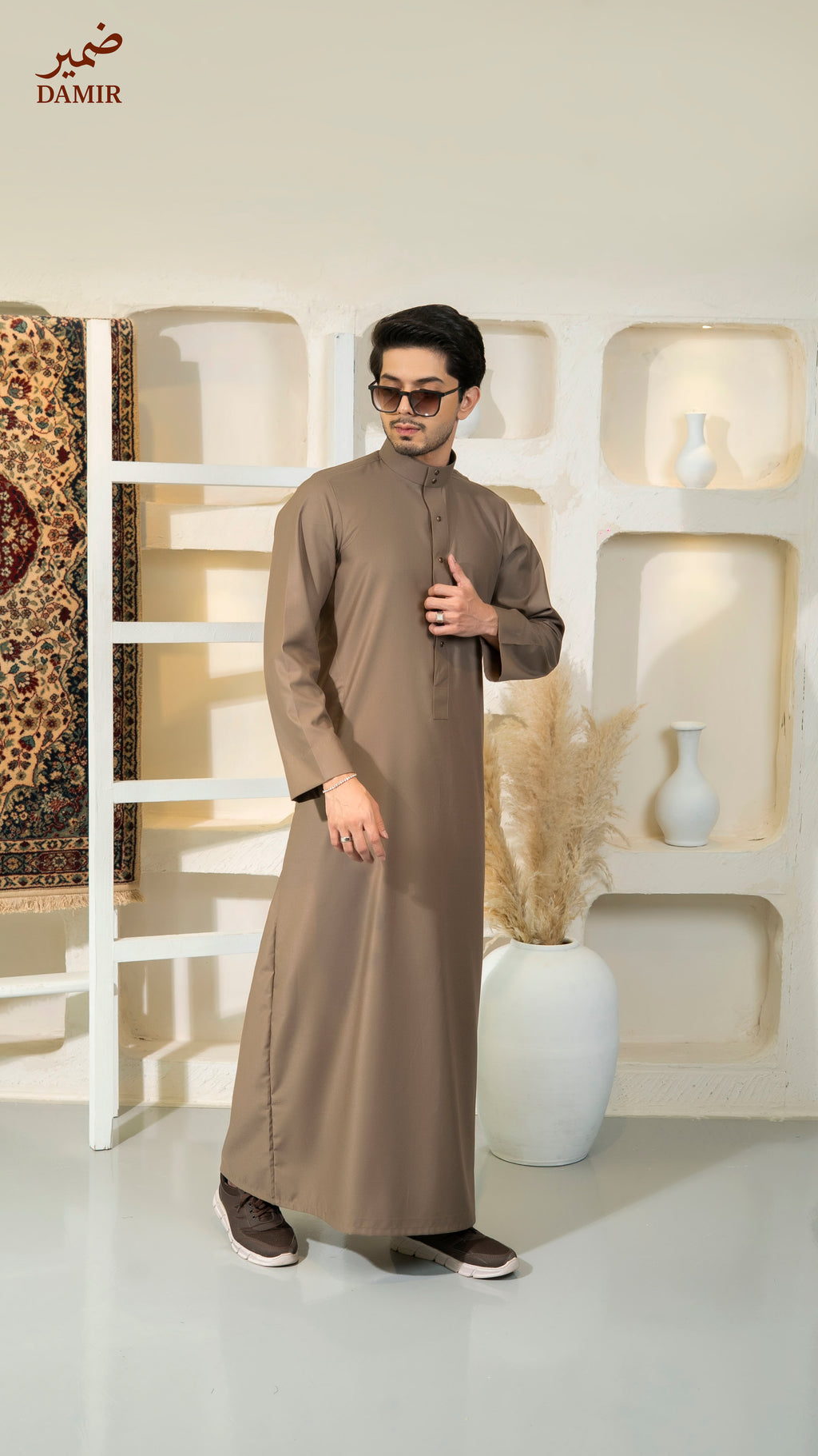 Man wearing a brown traditional outfit in a decorative room with 'DAMIR' branding.