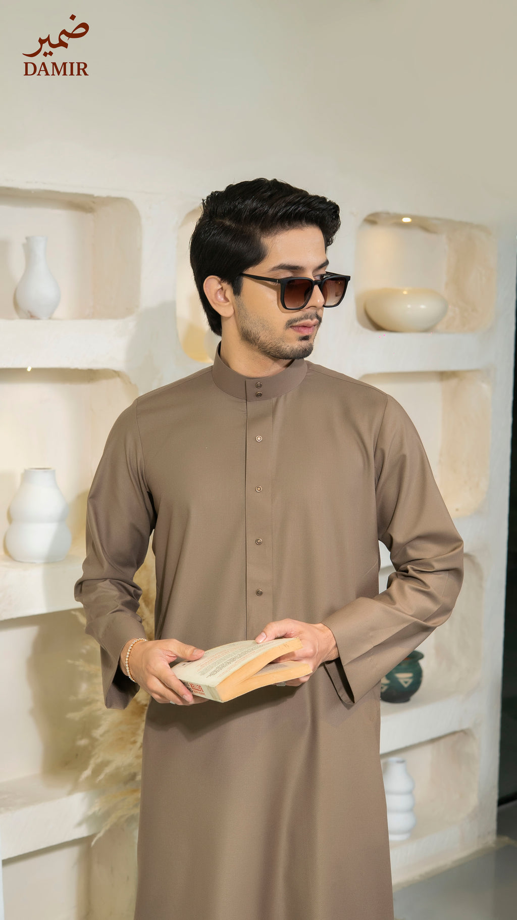 Man wearing a brown traditional outfit holding a book in a decorated room with 'DAMIR' branding.