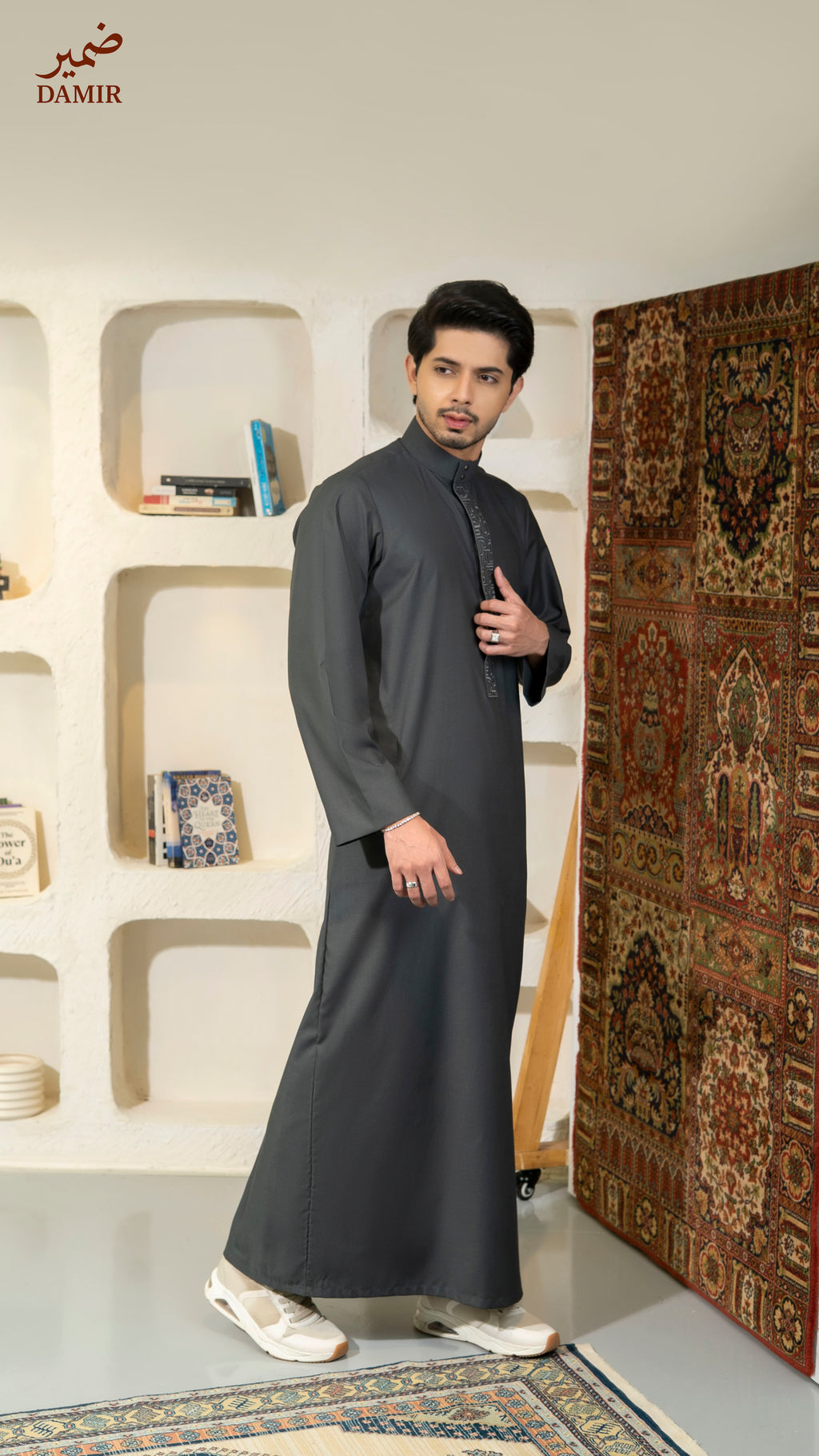 Man wearing a dark gray traditional outfit standing in a room with a decorative rug and shelves.