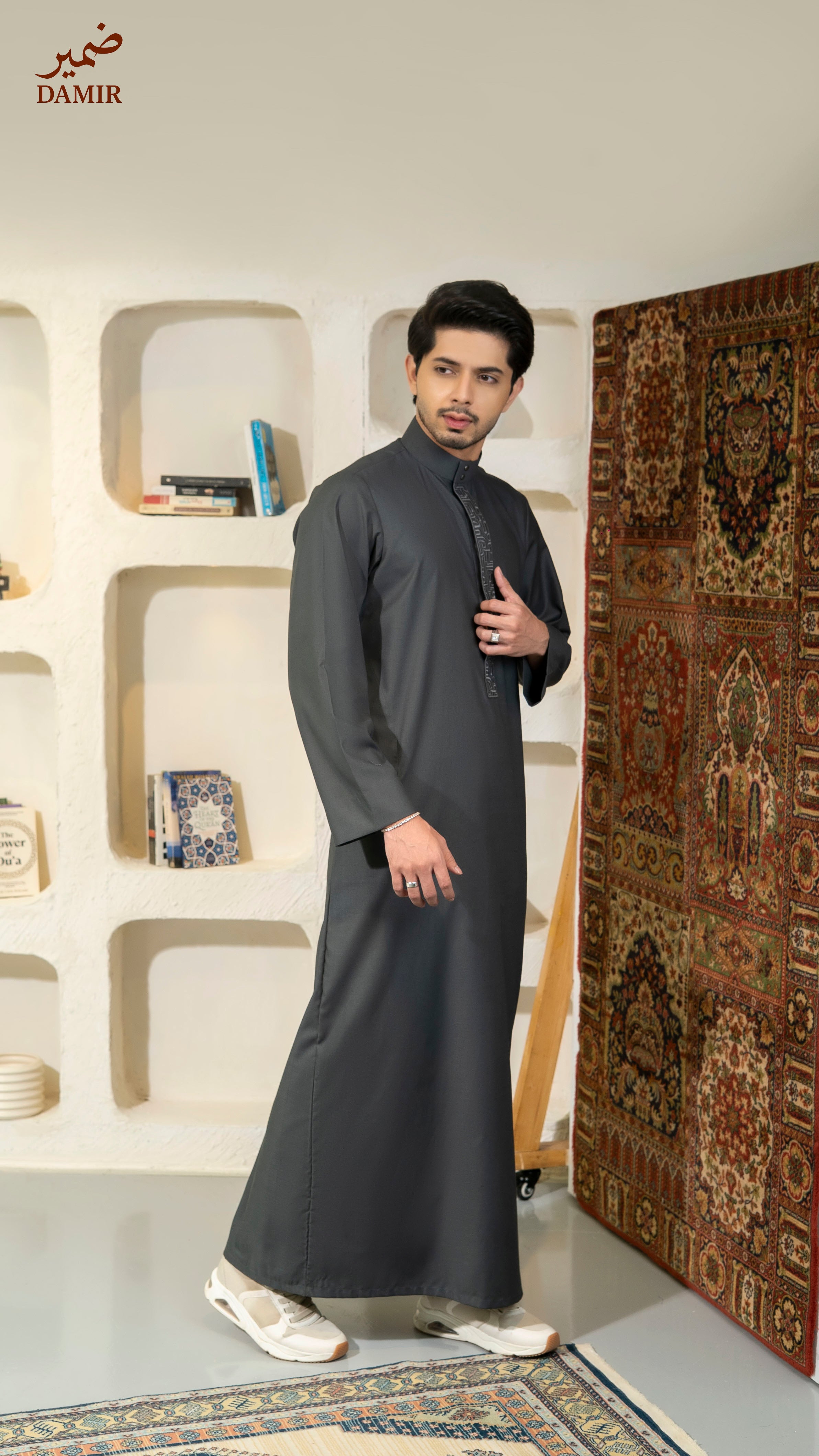 Man wearing a dark gray traditional outfit standing in a room with a decorative rug and shelves.