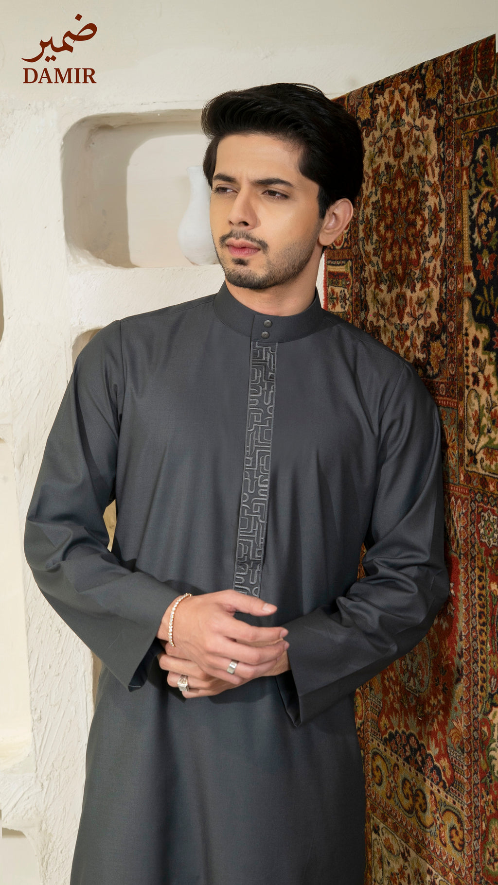 Man wearing a dark gray traditional outfit with intricate patterns, standing against a textured wall.